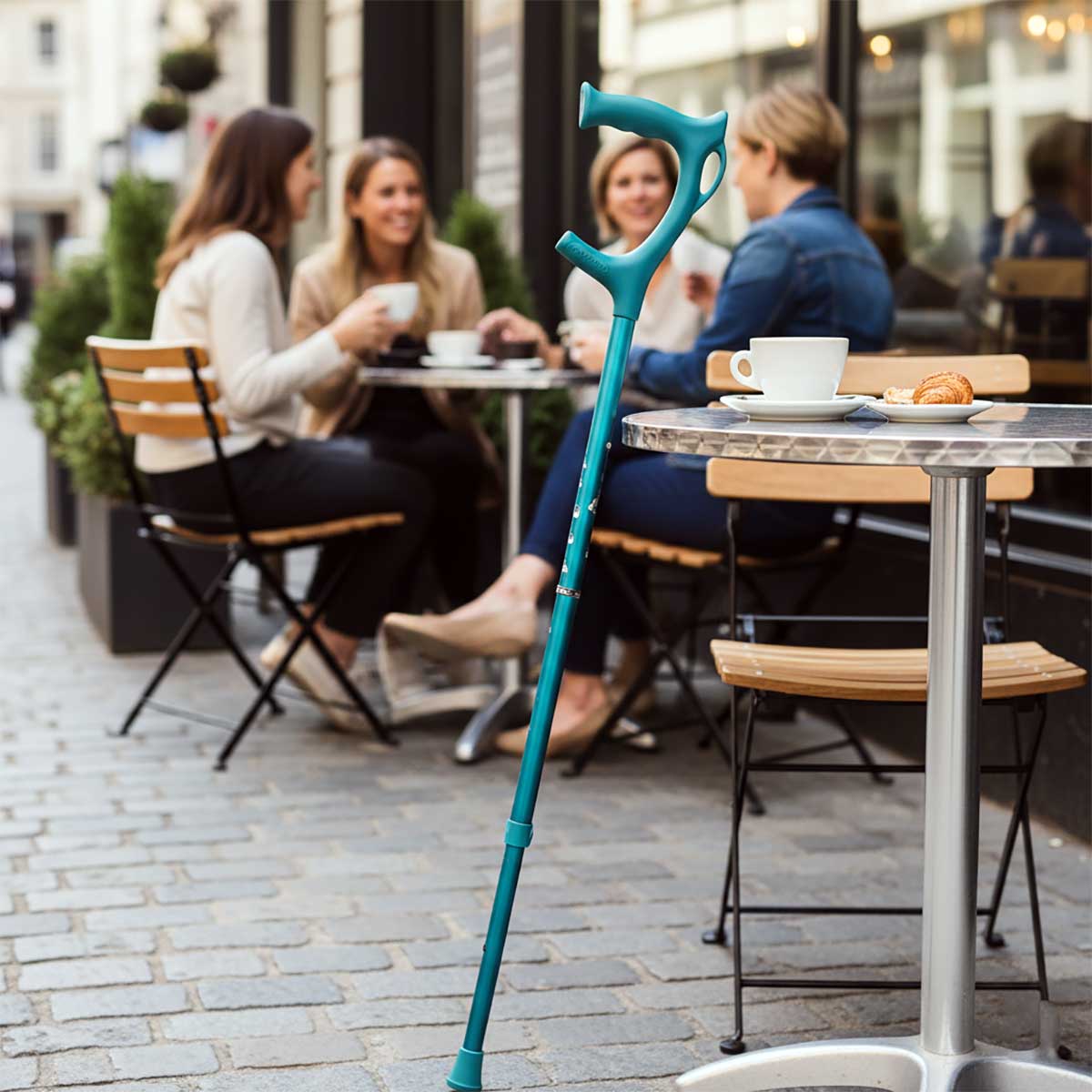 Cool Crutches folding walking stick in teal leaning against a cafe table, showing stylish design in a social setting