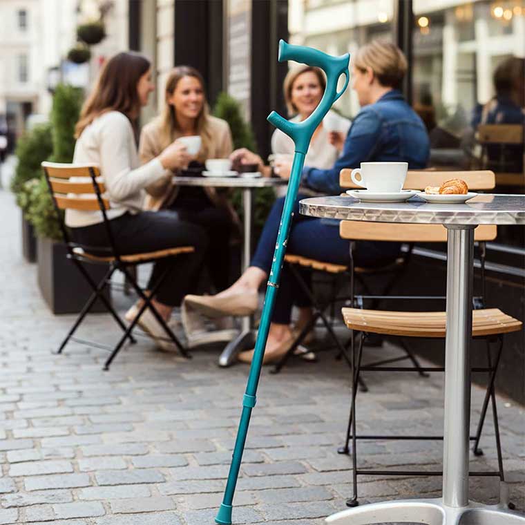 Cool Crutches folding walking stick in teal leaning against a cafe table, showing stylish design in a social setting