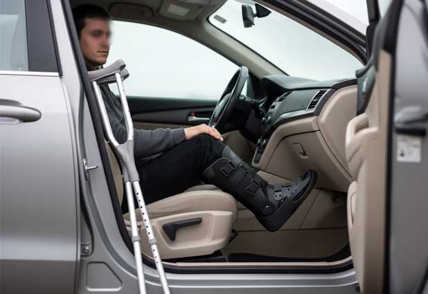 Person wearing a walking boot sitting in the driver’s seat with crutches beside the car.